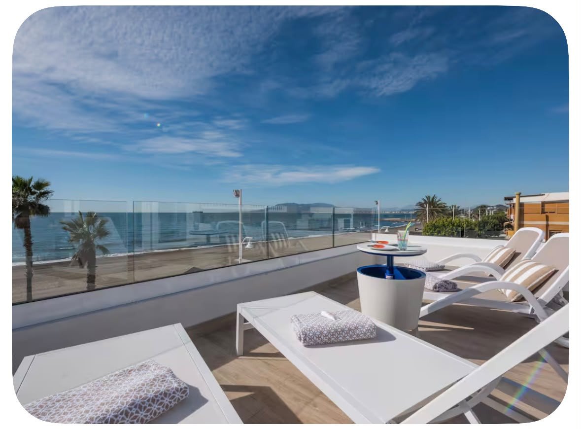Rooftop terrace with sun loungers, table, palm trees, and a clear view of the sea and horizon.