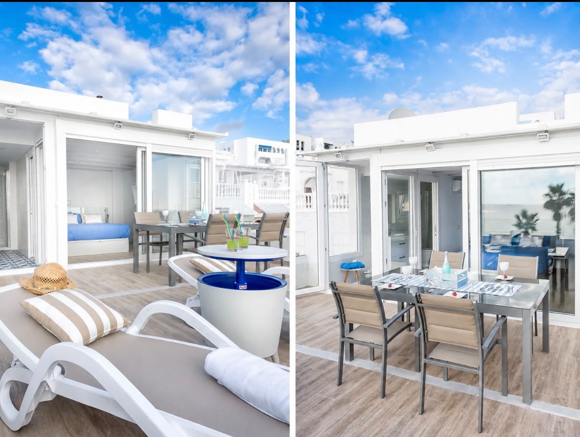 A sunny terrace with a bedroom visible inside, an outdoor dining area, and a glimpse of the sea beyond.