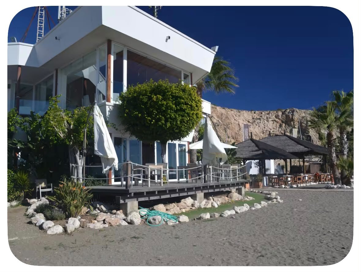 Modern white building with a wooden deck and outdoor seating area on a sandy beach with palm trees and cliffs.