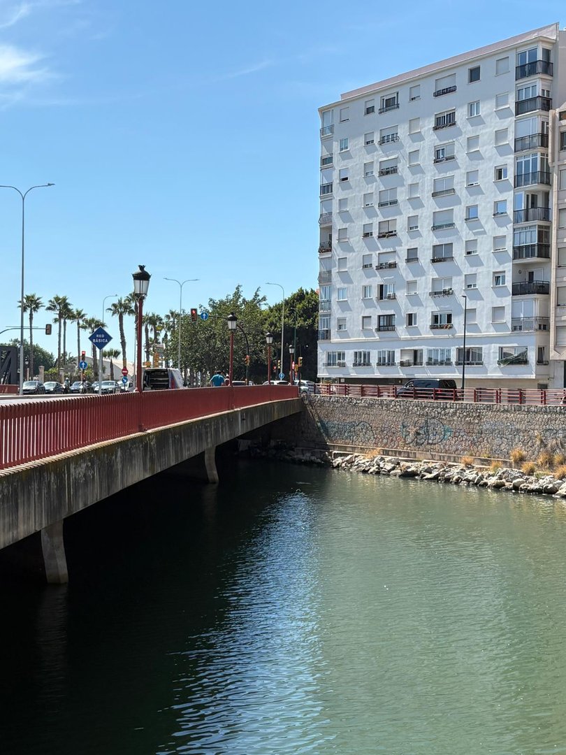 A bridge crosses a river next to a tall building with many windows, under a clear blue sky.