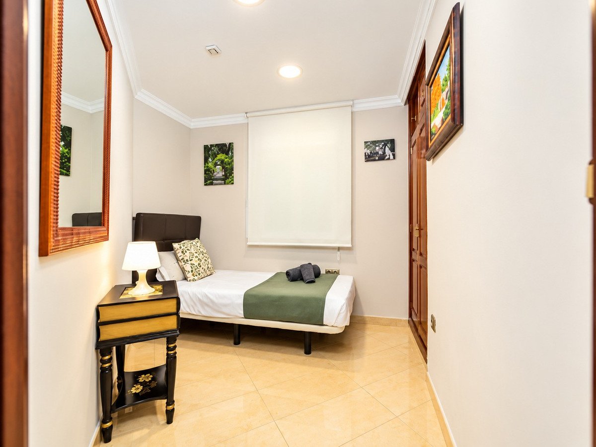 A single bed with green blanket in a minimalist room, featuring a bedside table, a mirror, and artwork on the walls.