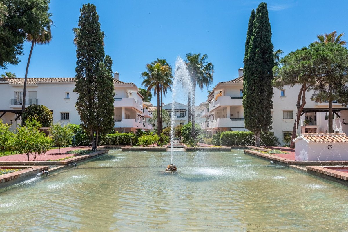 A sunny courtyard with a central fountain, palm trees, and white apartment buildings.