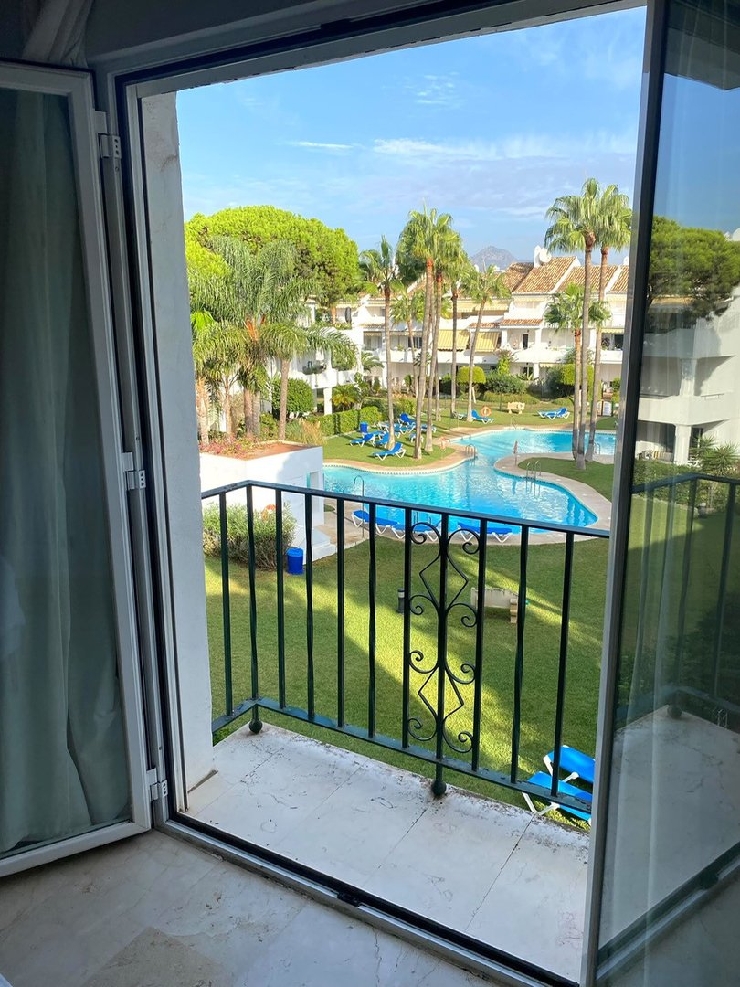 View from a balcony overlooking a swimming pool surrounded by palm trees and manicured lawns. Sunny day with blue sky.