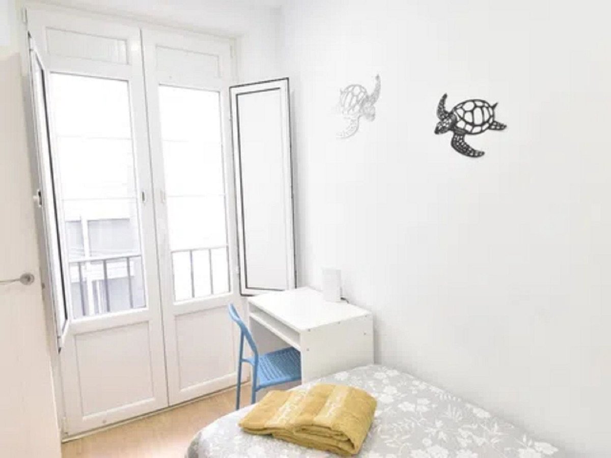 A small bedroom with a white desk, blue chair, and a bed with yellow blankets. Features a balcony door with shutters.