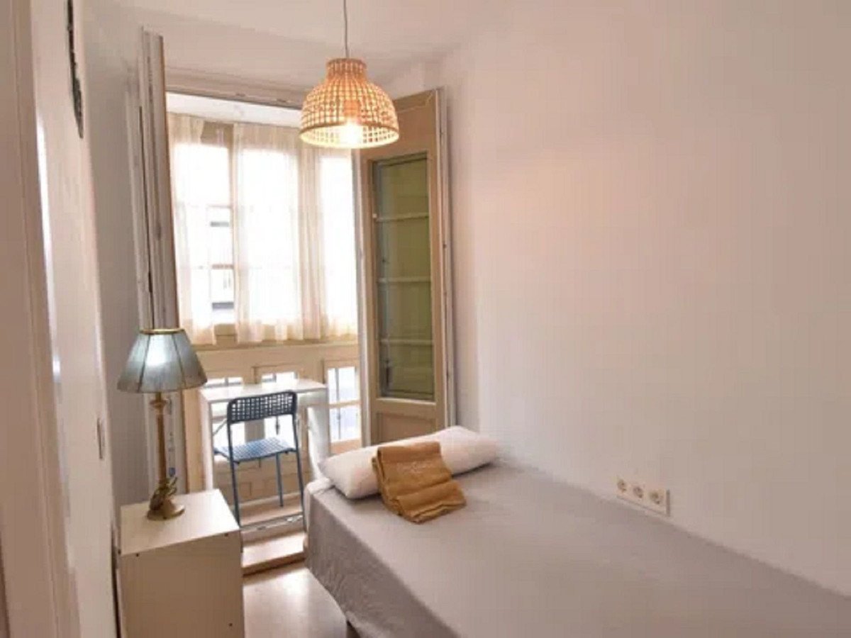 A small bedroom with a single bed, a bedside table, a lamp, and a door leading to a balcony with a table and chair.