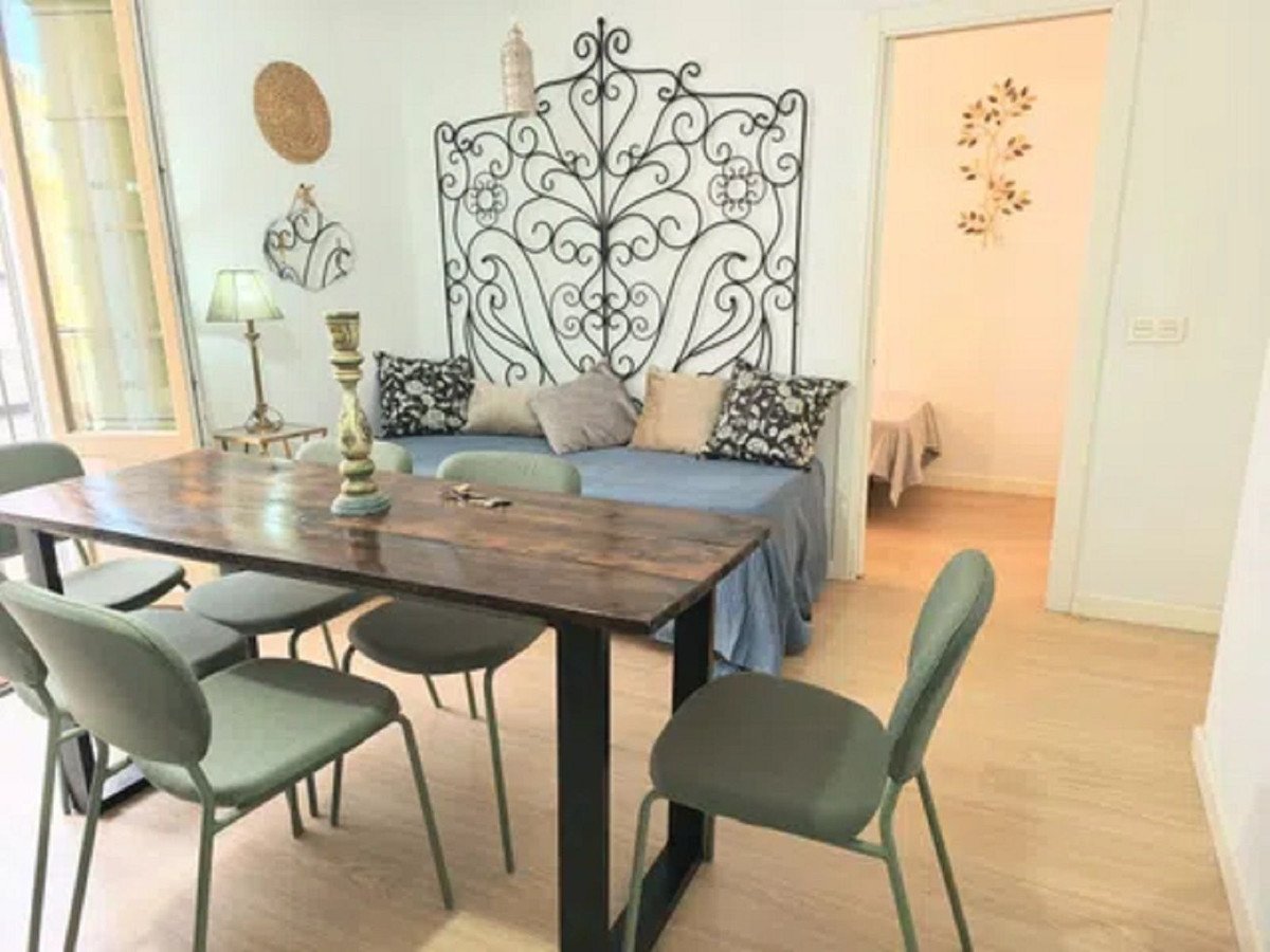 A wooden dining table with chairs is in front of a daybed with decorative pillows and an ornate headboard. A doorway leads to another room.