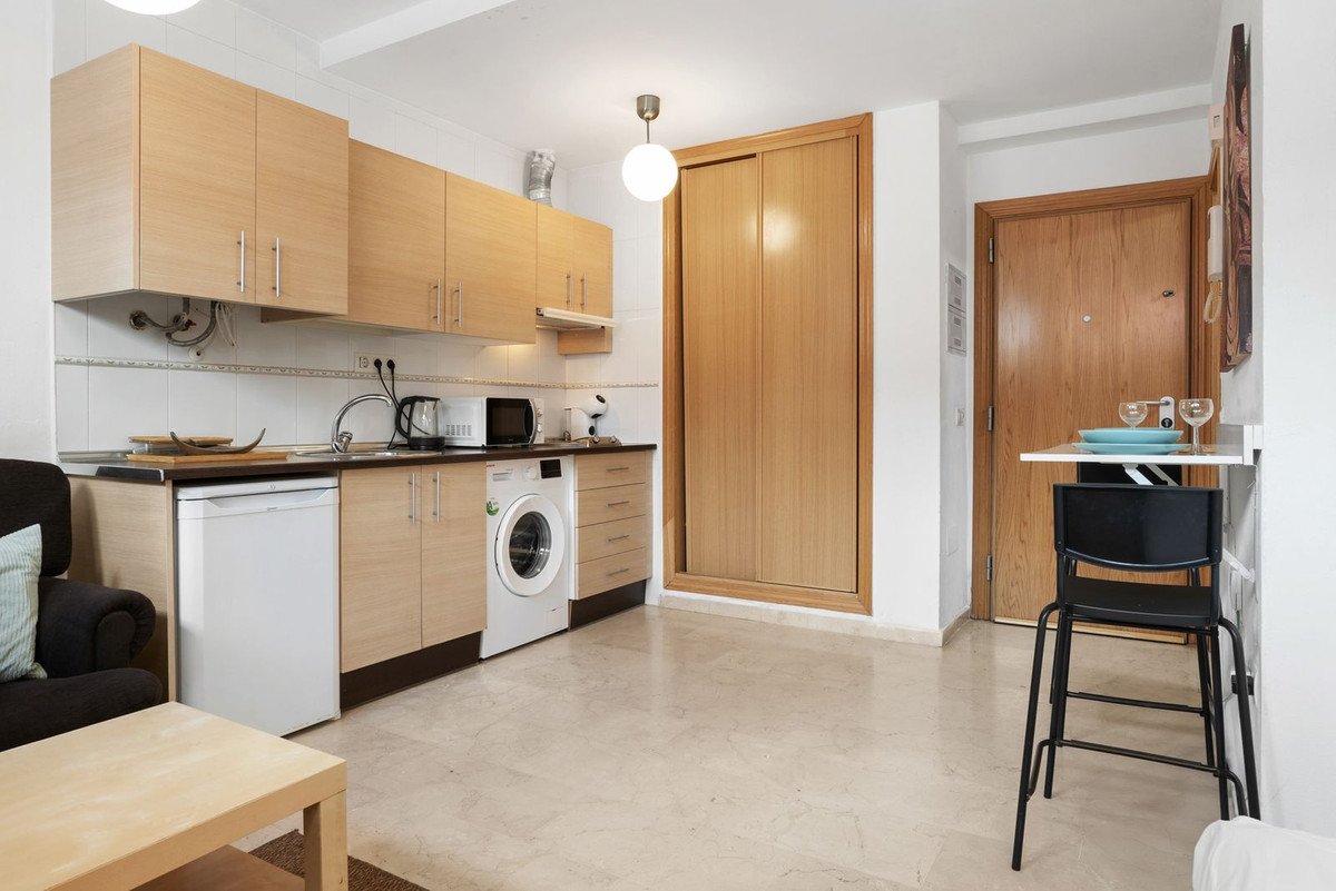A compact kitchen with wood cabinets, a washing machine, refrigerator, and a small dining area with a stool.