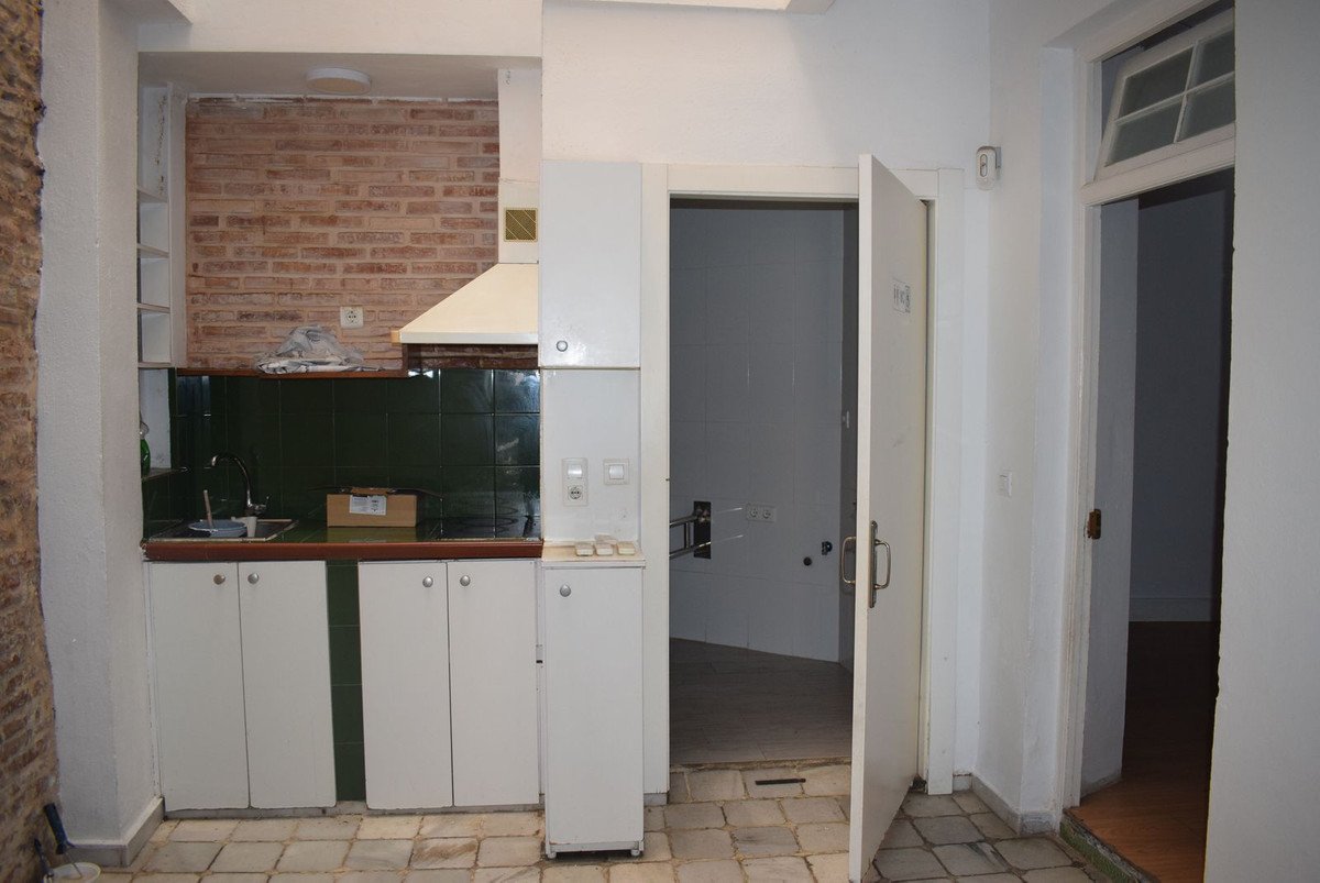 A compact kitchen area with white cabinets, dark green tiles, and a brick wall background. Tiled floor.