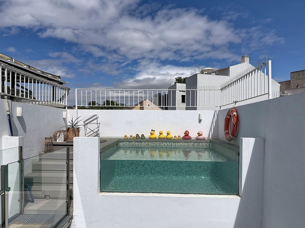 A modern rooftop pool with a glass bottom, decorated with rubber ducks and a life preserver, under a cloudy blue sky.