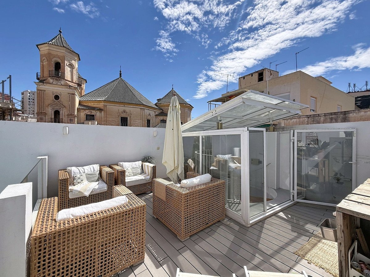 Rooftop terrace with wicker furniture and a glass enclosure, overlooking historic church buildings under a blue sky with clouds.