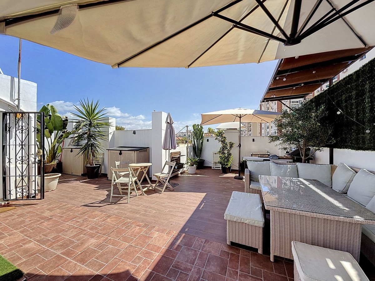 Spacious rooftop terrace with dining set, lounge area, umbrellas, and lush plants under a clear blue sky.