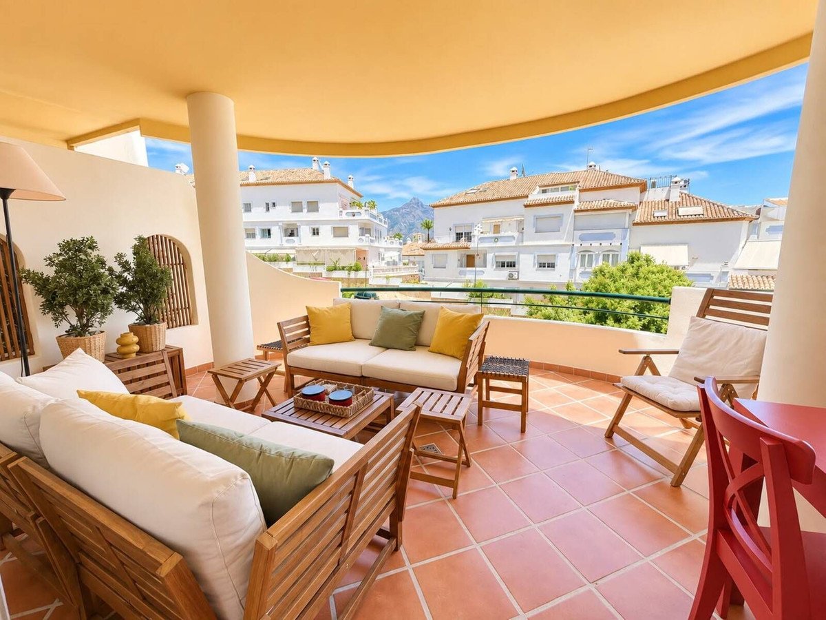 Spacious terrace with wooden seating area, coffee tables, and views of white buildings and blue sky.