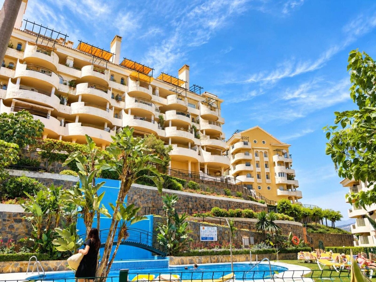 A sunny view of a multi-story apartment complex with balconies, a swimming pool, and lush greenery.