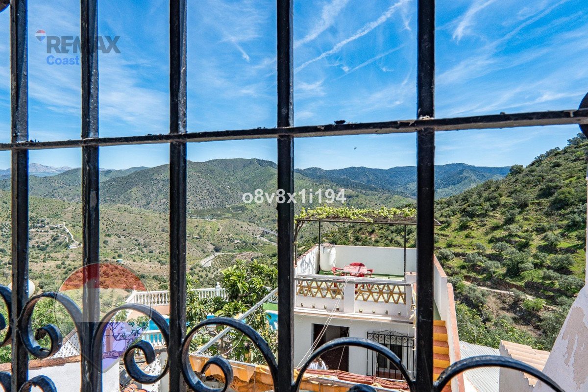 View of rolling green hills and mountains through decorative bars, with a tiled terrace and red chairs visible.