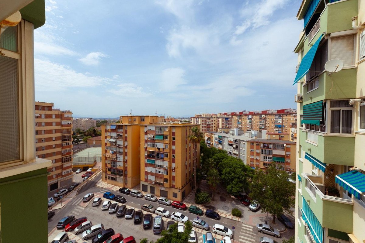 View from a balcony overlooking a city street with parked cars and apartment buildings under a cloudy sky.