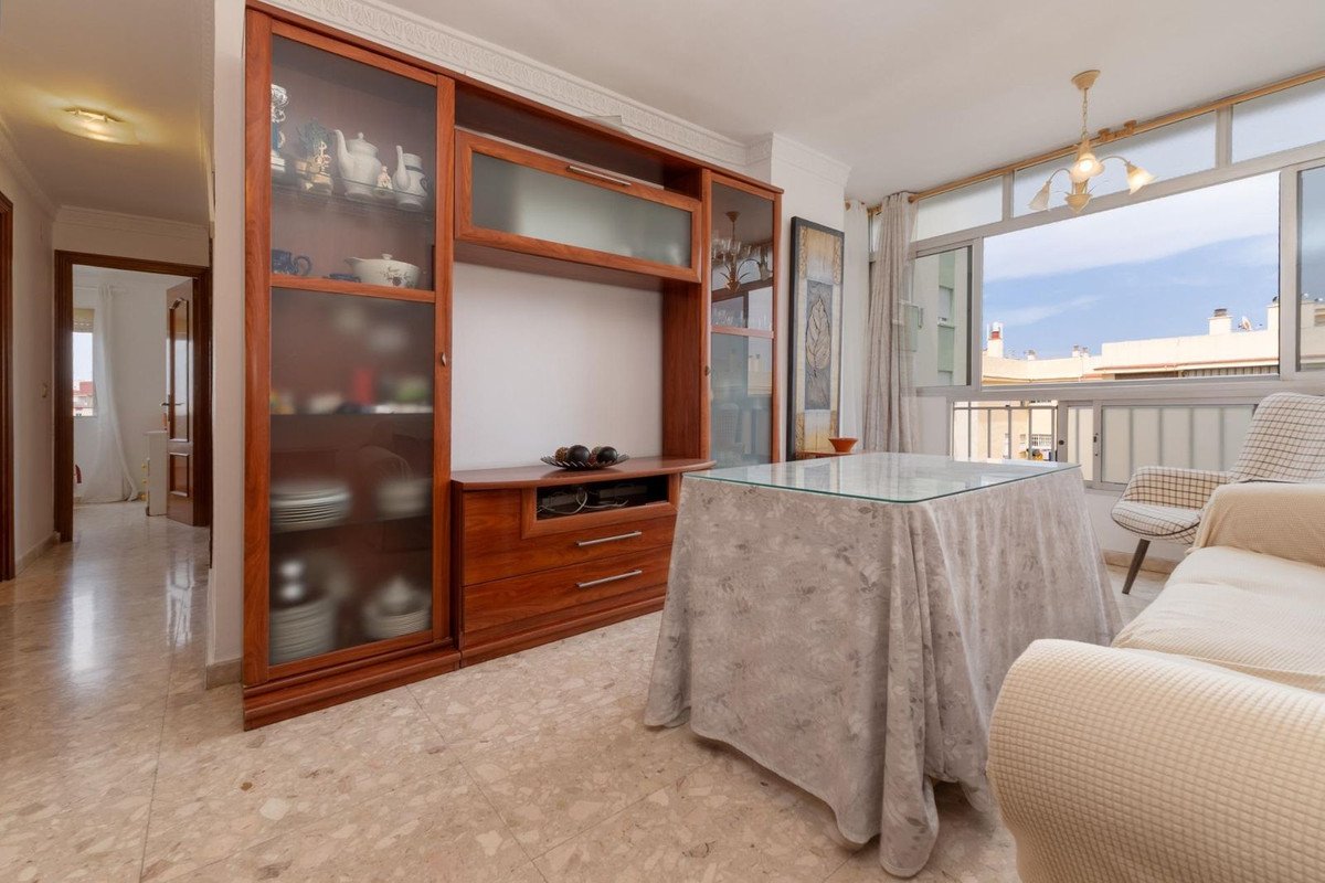 Living room with wooden cabinets, covered table, and balcony overlooking rooftops under a bright blue sky.