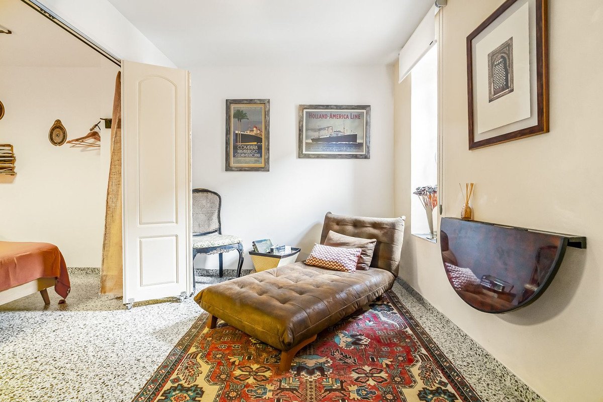 A brown chaise lounge sits on an ornate rug in a room with vintage posters and a small folding table by the window.