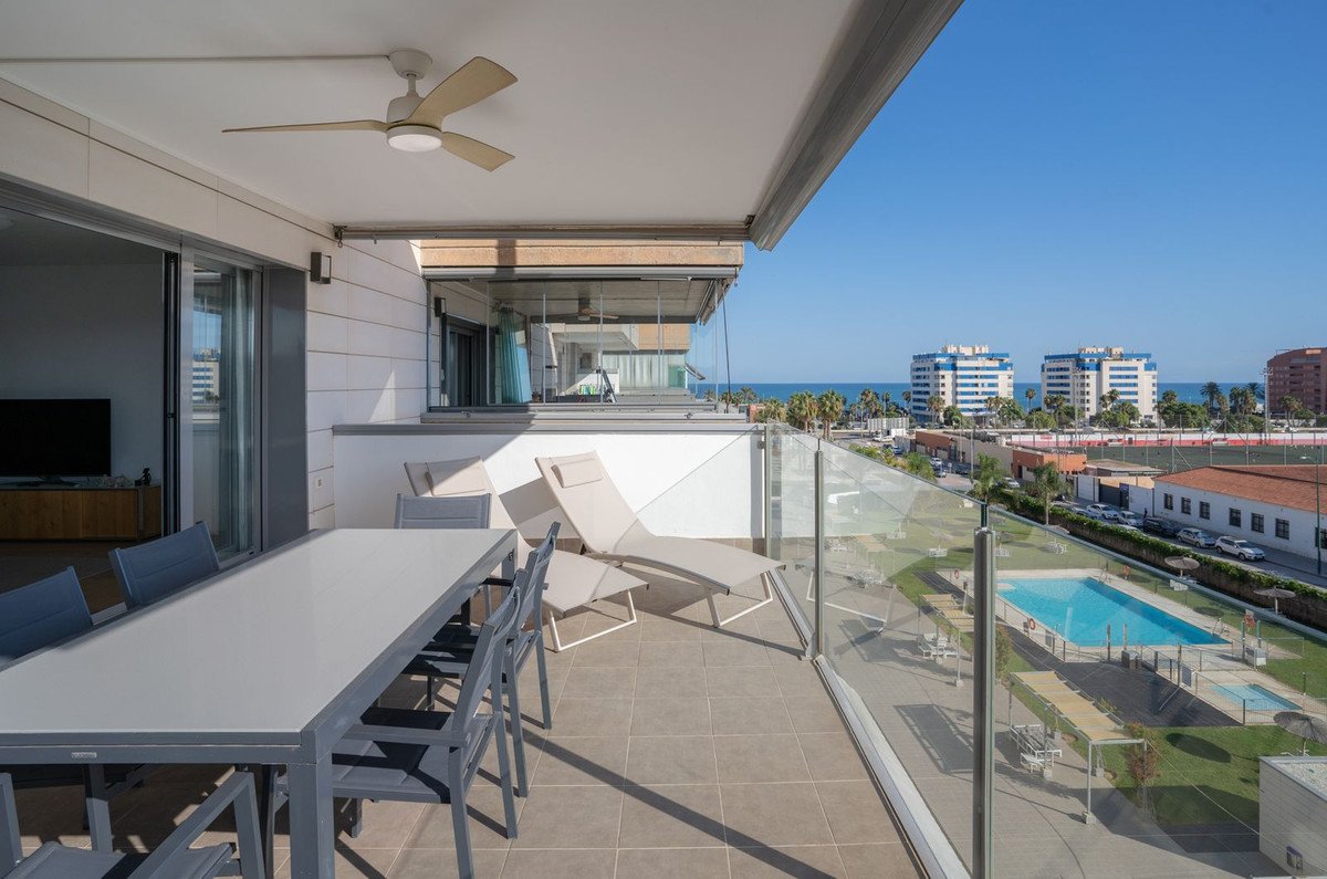 Modern balcony with dining set and lounge chairs overlooking a pool and the sea on a sunny day.
