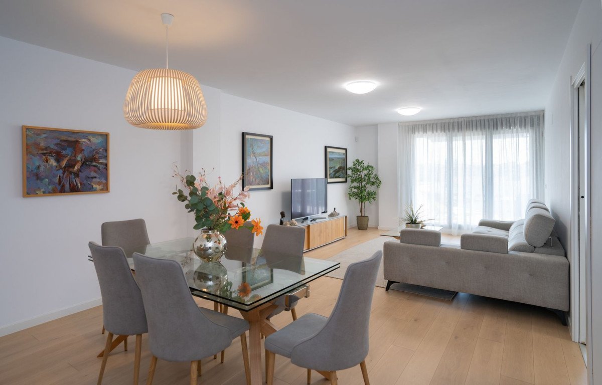 Modern dining and living area with grey upholstered chairs, glass dining table, sofa, TV, and large window with sheer curtains.