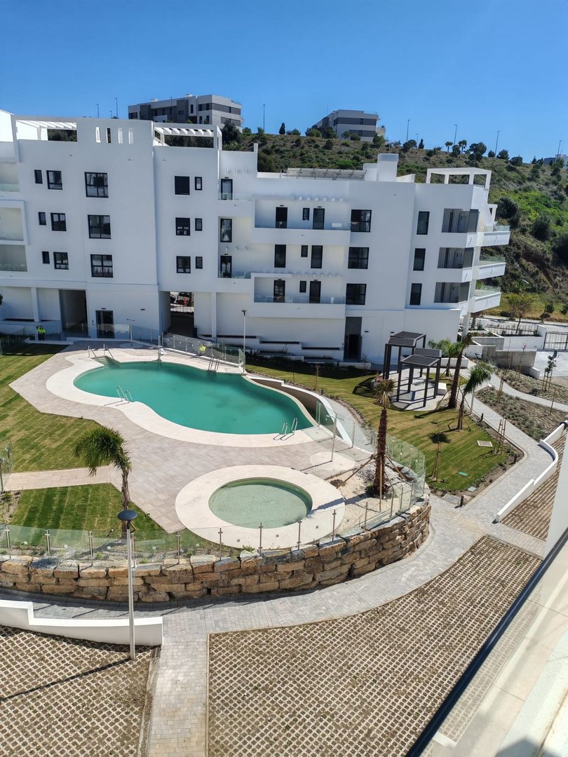 Modern white apartment building with balconies overlooking a curved swimming pool and jacuzzi surrounded by landscaped gardens and pathways.