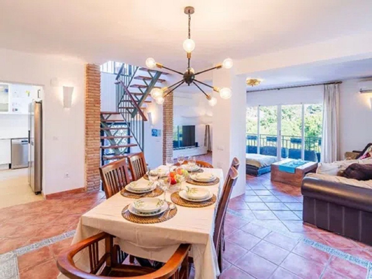 A spacious dining room with a large table, leading to a living area with a sofa and a view of lush greenery through the balcony doors.