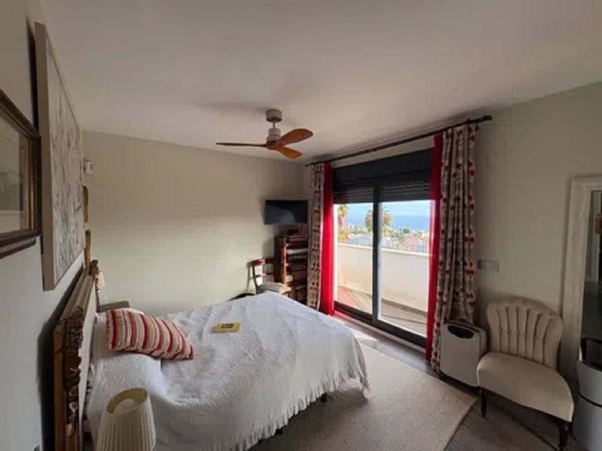 A bedroom with a ceiling fan, ocean view through sliding glass doors, and a white bedspread.