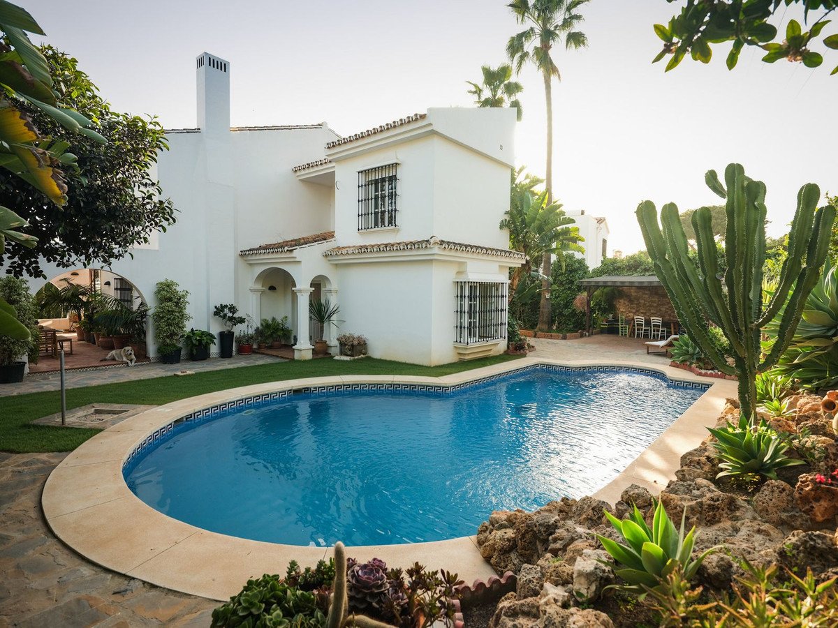 A white villa with a blue swimming pool, palm trees, and a cactus garden under a sunny sky.