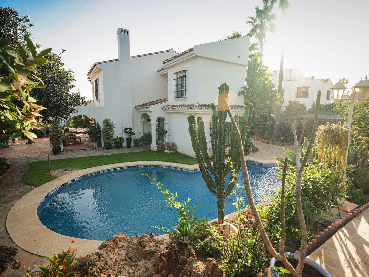 White villa with a kidney-shaped swimming pool and lush green garden, featuring large cacti and palm trees under a bright sunny sky.