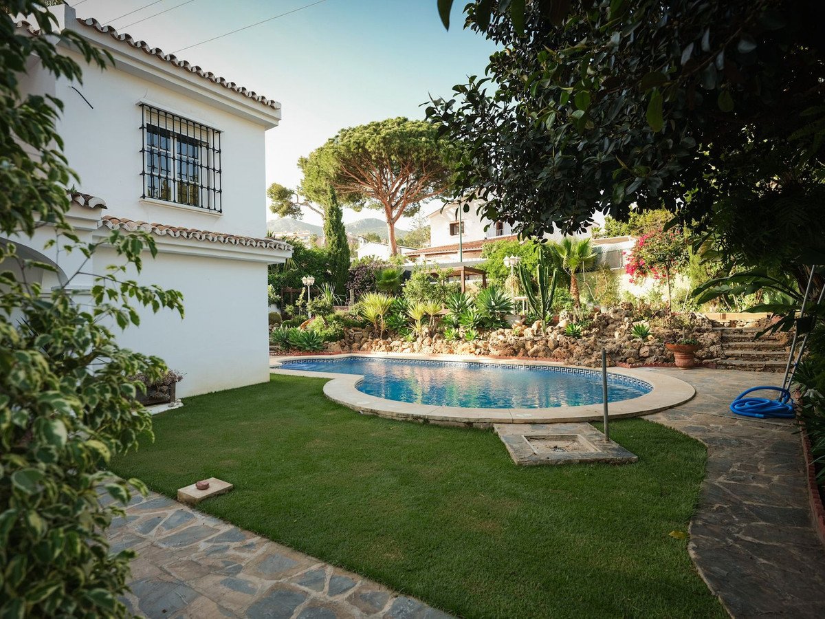 A white villa with a curved swimming pool, surrounded by a lush garden and mature trees, under a clear sky.