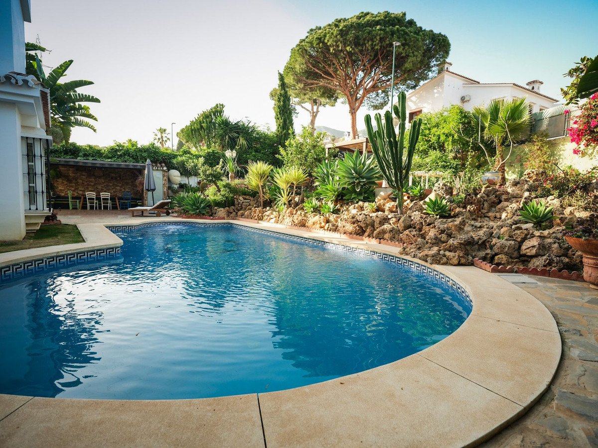 A bright, sunny day shows off a sparkling blue swimming pool surrounded by a rocky, xeriscape garden with mature trees and a white villa in the background.