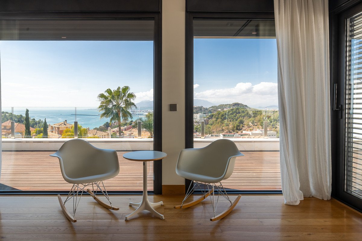 Two modern rocking chairs and a small table on a wooden floor, with large windows showing a sea view, palm tree, and hills.
