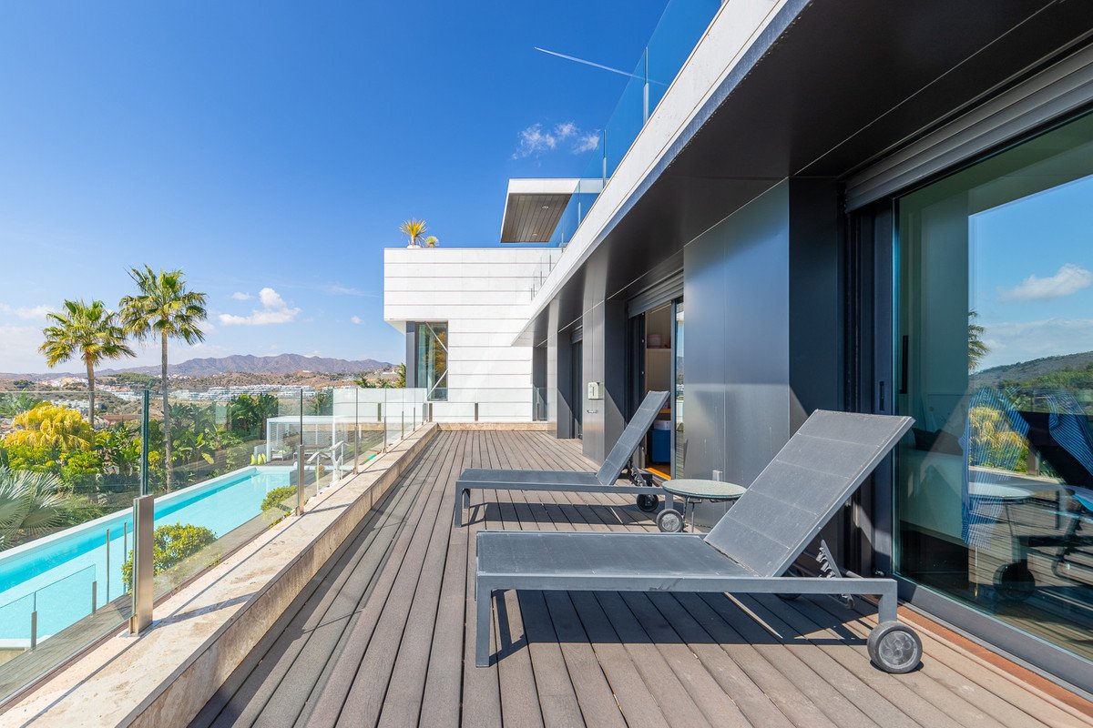 Modern terrace with lounge chairs, a long swimming pool, palm trees, and a view of the hills under a clear blue sky.