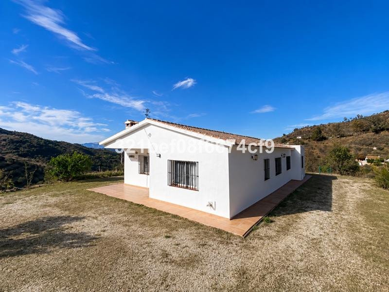 A white villa with a tiled terrace sits on a grassy hill with a clear blue sky and distant mountains.