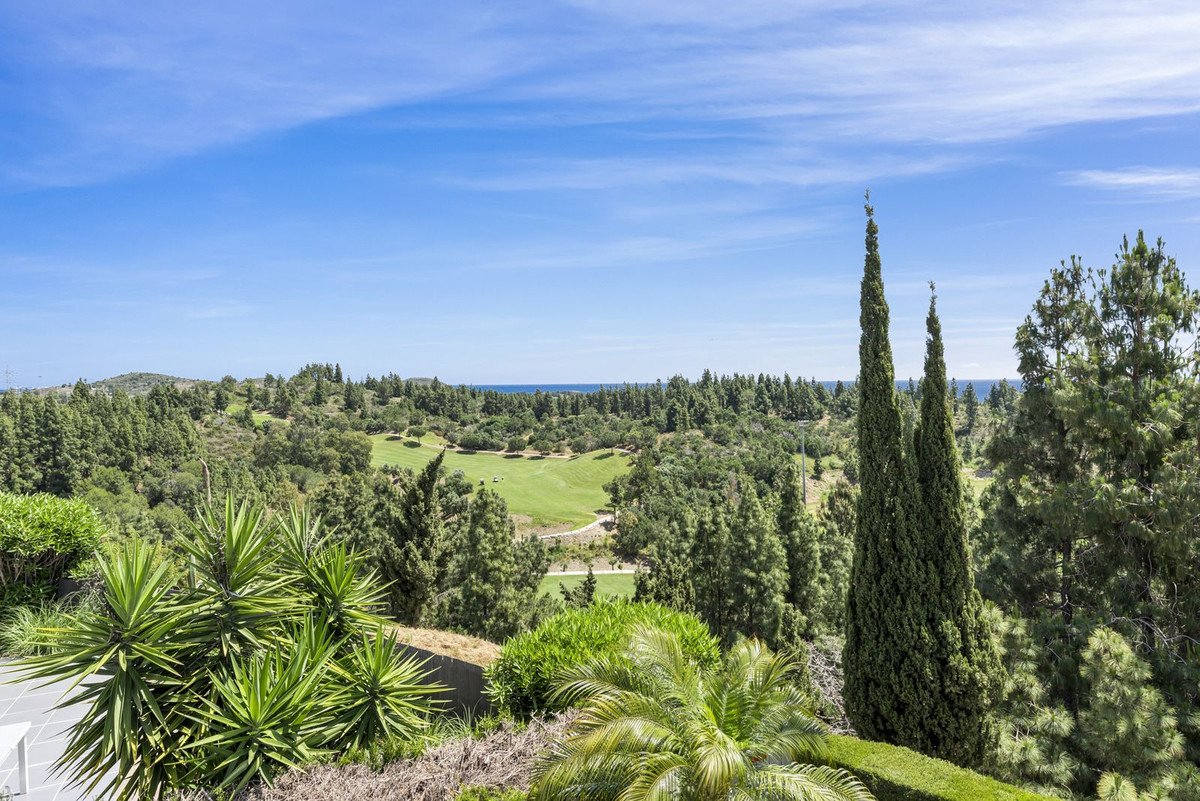 Expansive green golf course with manicured fairways, lush trees, and a distant view of the sea under a clear blue sky.