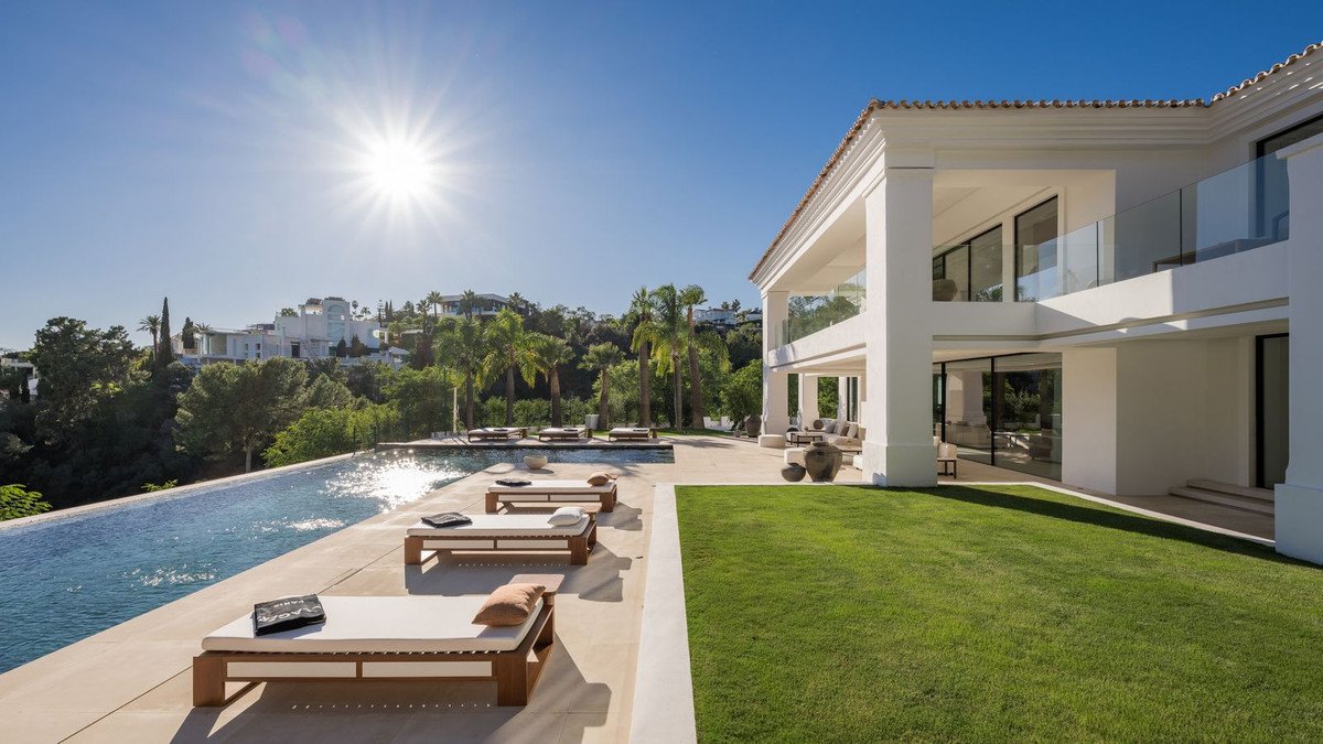 Modern villa with an infinity pool, lounge chairs, green lawn, and a balcony overlooking lush hills under a bright sun.