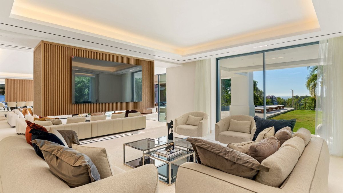Spacious modern living room with large sectional sofa, sectional seating, a wall-mounted TV, and panoramic windows overlooking a green lawn and sea.