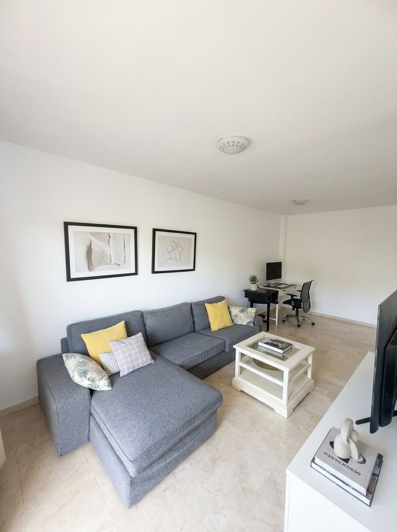 A modern living room featuring a grey sectional sofa, coffee table, desk with computer, and minimalist artwork on the wall.