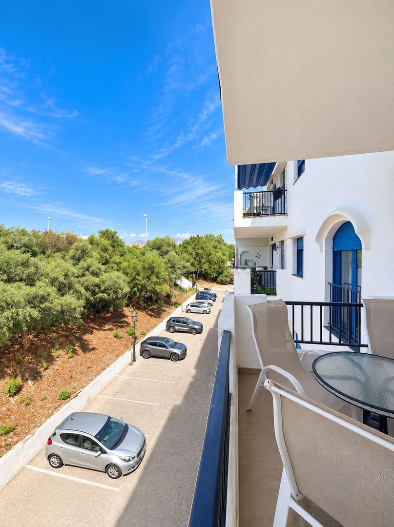 View from a balcony overlooking a parking area with cars, surrounded by lush greenery and trees under a blue sky.