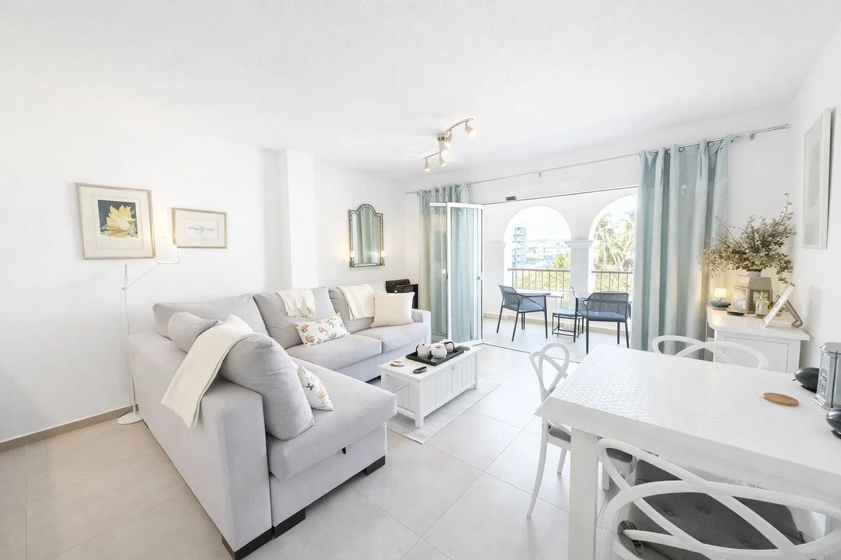 Bright living and dining area with a grey sectional sofa, white dining table, and access to a balcony with outdoor seating.