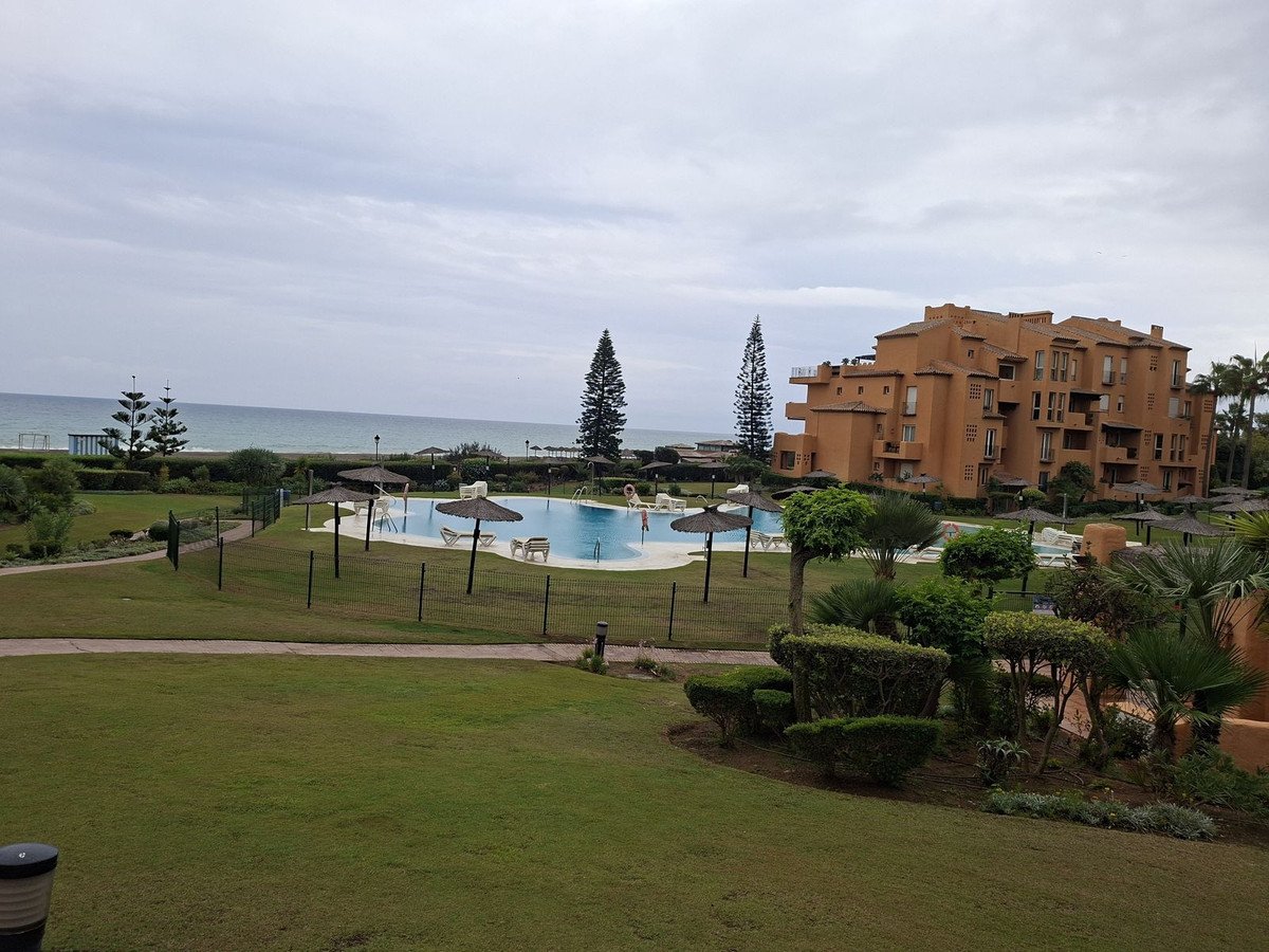 Apartment complex with a swimming pool, lush gardens, and a view of the sea under a cloudy sky.