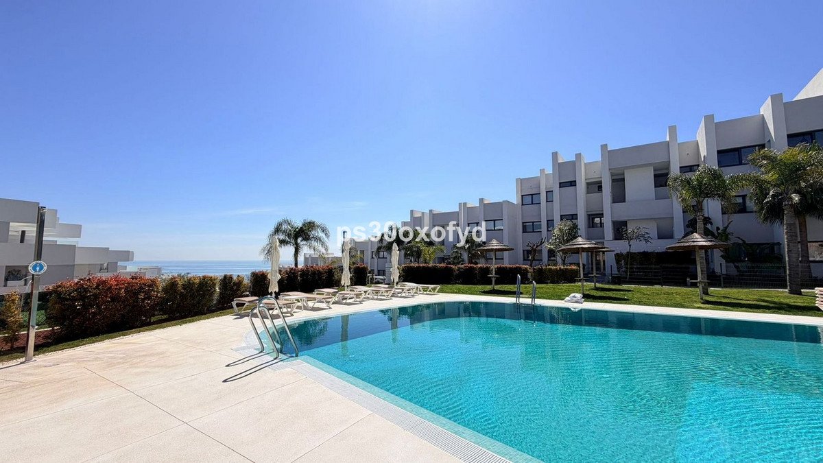 Communal swimming pool with sun loungers and umbrellas, overlooking the sea and modern apartment buildings.