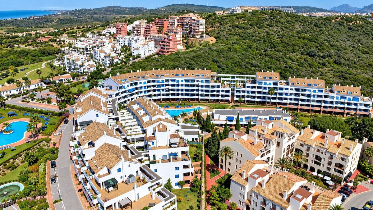 Aerial view of modern apartment complex with swimming pools, lush landscaping, and distant sea views under a clear blue sky.