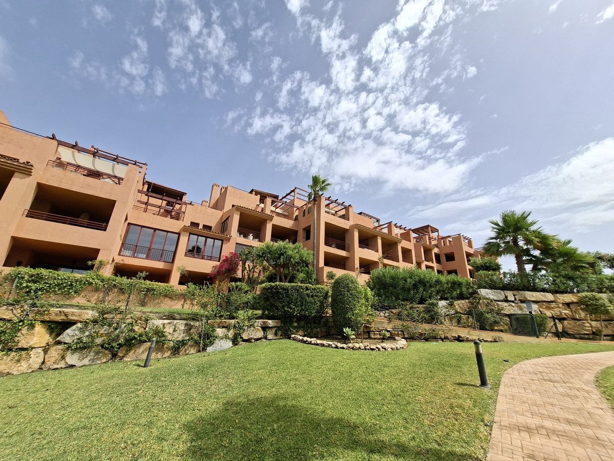 Terraced apartments with balconies and lush greenery, overlooking a paved walkway and garden under a partly cloudy sky.