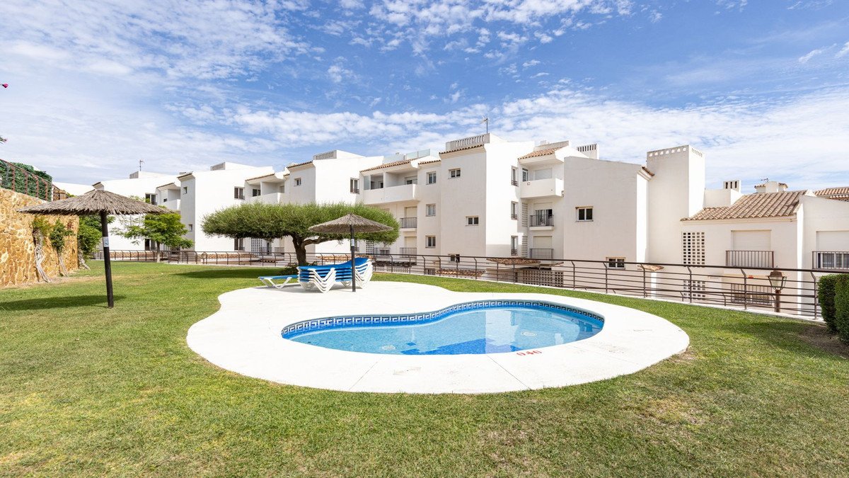 A kidney-shaped swimming pool on a green lawn with lounge chairs and umbrellas, in front of white apartment buildings.