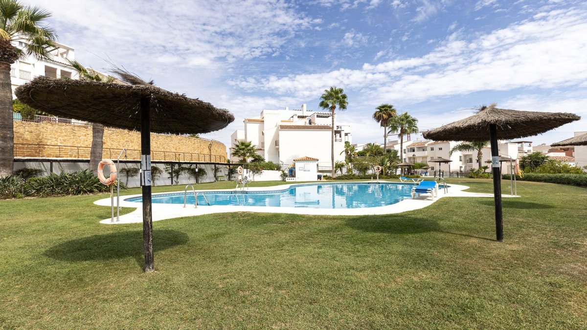 A resort swimming pool area with lounge chairs and thatched umbrellas, surrounded by palm trees and apartment buildings.