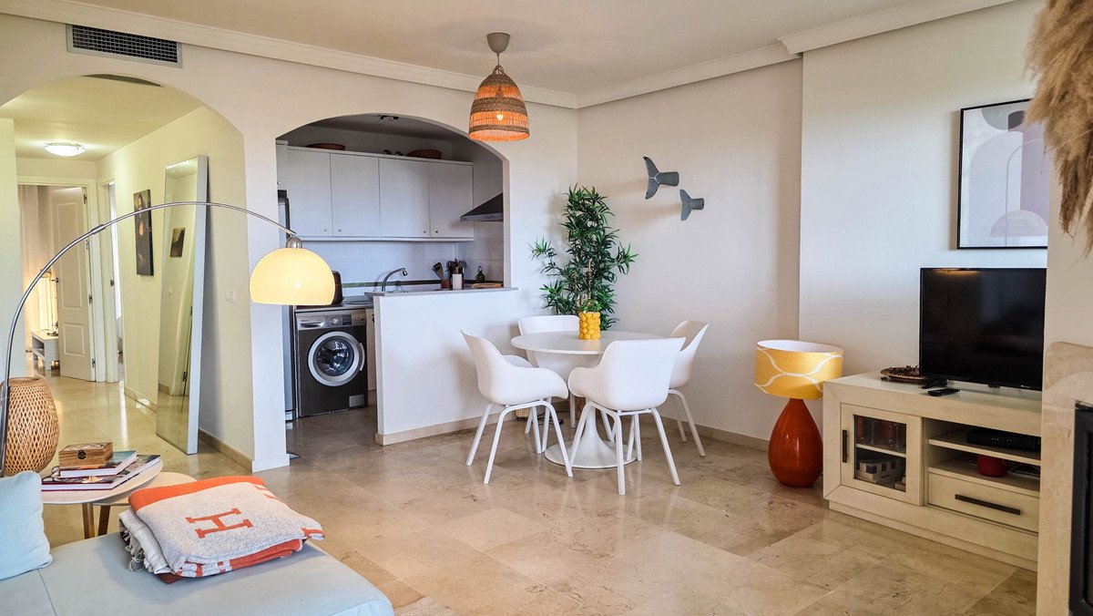 Open-plan living, dining, and kitchen area with modern white furniture, TV, and natural light.