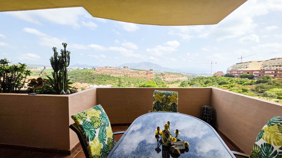 Terrace with a table and chairs, overlooking a green landscape with hills and buildings under a cloudy sky.