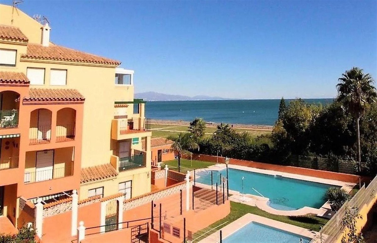 Apartment building with balconies overlooking the sea, swimming pools, and palm trees under a clear blue sky.
