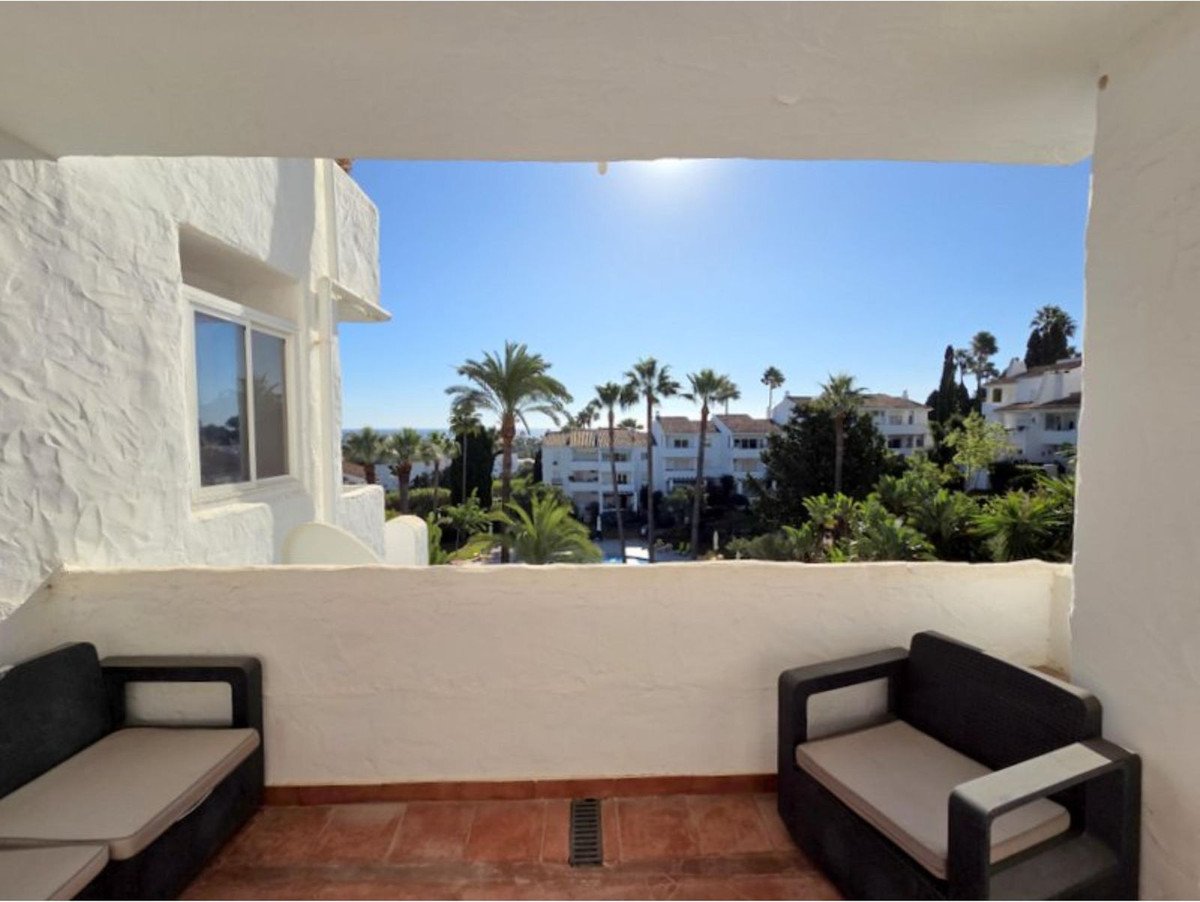 Balcony with outdoor seating offers a view of white buildings, palm trees, and the sea on a sunny day.