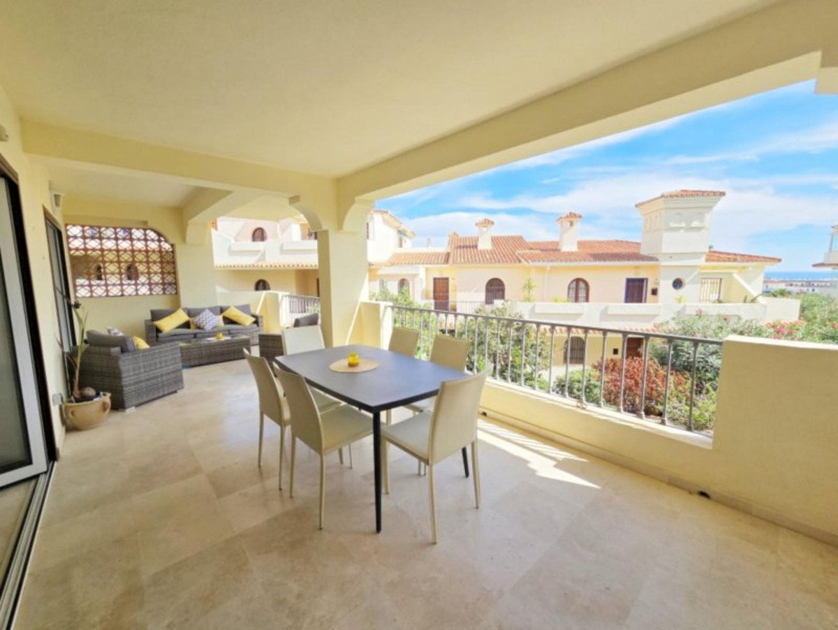 A covered terrace with dining table, seating area, and a view of Mediterranean-style houses and lush greenery.